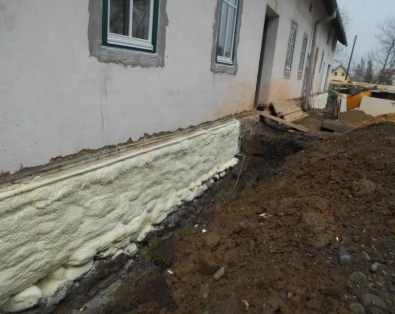 Bílá pěnová izolace aplikovaná na základnu domu před zasypáním.