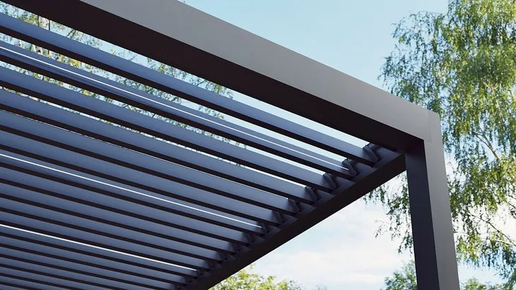 Moderní bioklimatická pergola s nastavitelnými lamelami na pozadí zelených stromů.