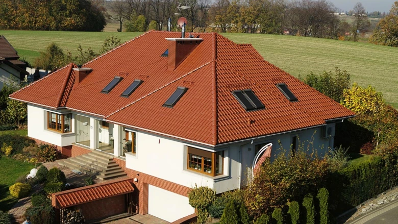 Moderní dům s polovalbovou i valbovou střechou na pozadí zelené krajiny.