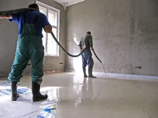 Dva dělníci nalévají betonový potěr do nehotové místnosti.