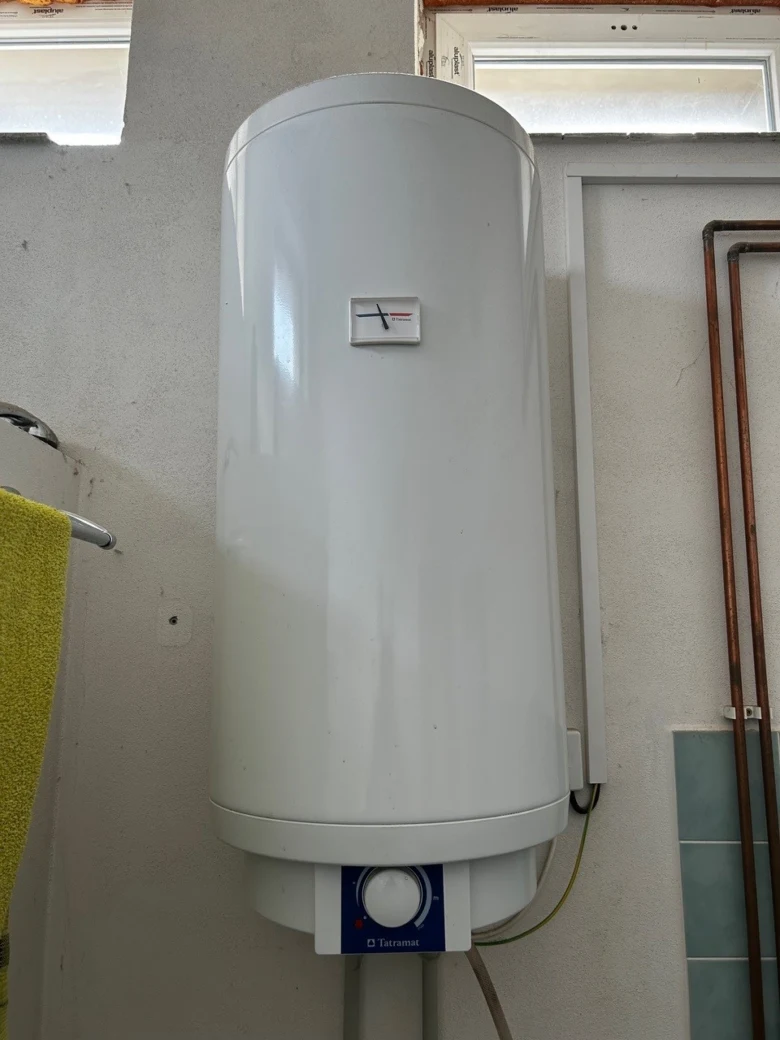 Bílý elektrický bojler na stěně vedle měděných potrubí.