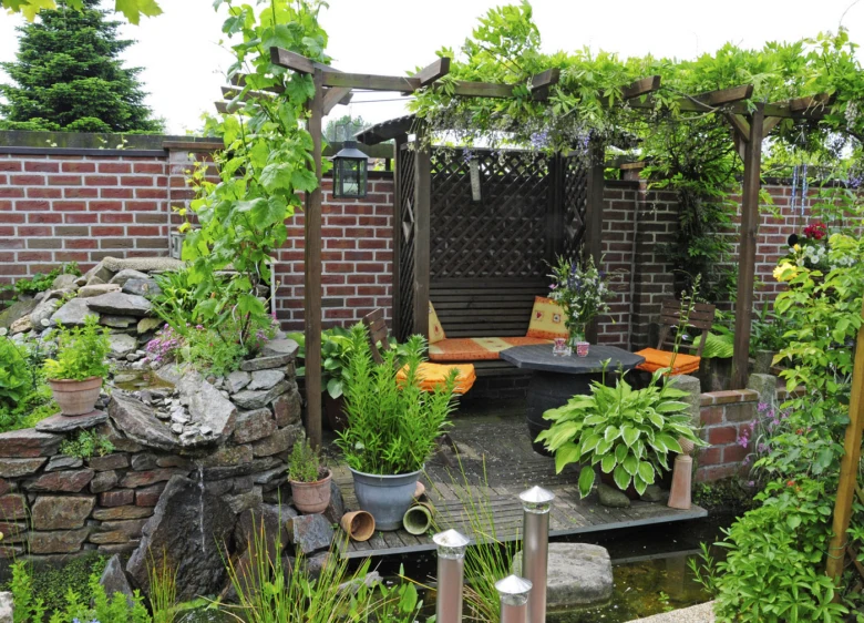 Malá dřevěná pergola na zahradě s posezením a vodním prvkem.
