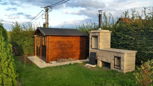 Dřevěná chata vedle kamenného grilu v zahradním pozadí.