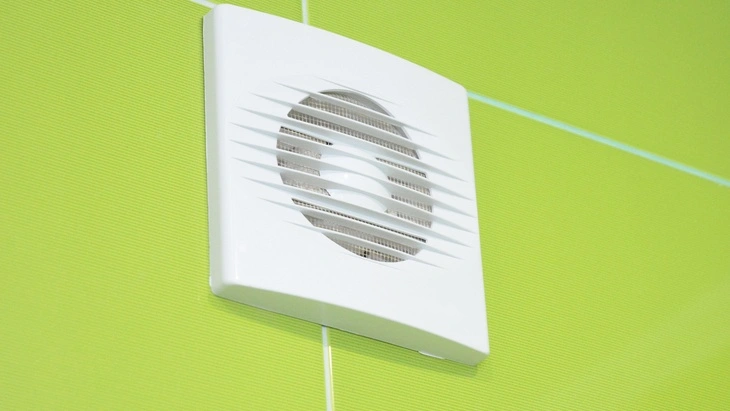 Bílý ventilátor pro odvětrání koupelny na zelené stěně.