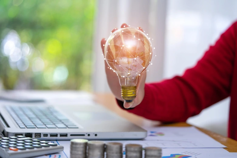 Ruka drží rozsvícenou žárovku s digitální mřížkou nad pracovním stolem