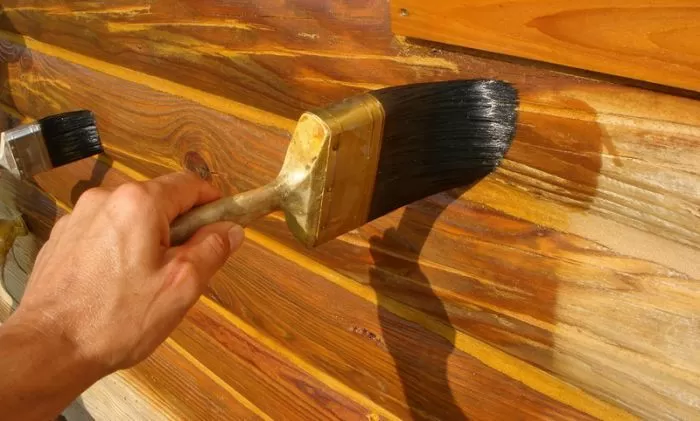 Ruka nanáší venkovní lazuru na dřevěné obložení štětcem.
