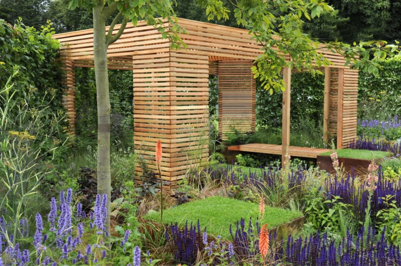 Jednoduchá dřevěná pergola v zahradě s kvetoucími rostlinami.