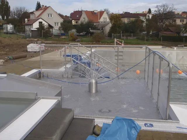 Nově budovaný venkovní bazén s fontánou na ploše obklopený zábradlím.