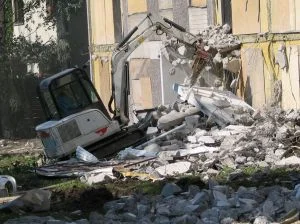 Bílý bagr rozebírá zdi budovy při demolici.
