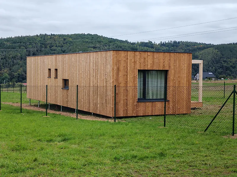 Moderní dřevěný modulární dům stojí na trávníku před lesními kopci.