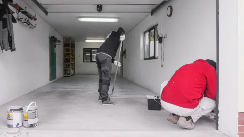 Dva lidé malují betonovou podlahu v garáži válečkem.