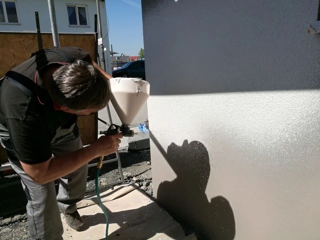 Pracovník aplikuje stříkanou omítku na fasádu pomocí pistole.