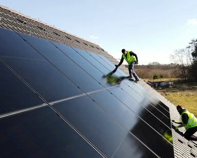 Dva dělníči čistí fotovoltaické panely na pultové střechě.