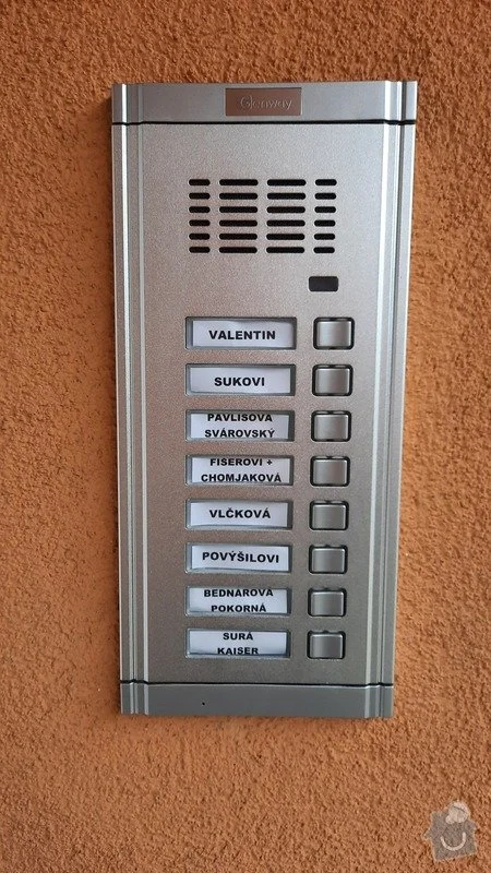 Stříbrný panel domovního zvonku s tlačítky pro jednotlivé byty.